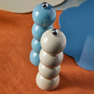 Two stacked Addison Ross London Bobbin Salt or Pepper Grinders, one in pastel blue and the other in off-white, stand on a textured burnt orange surface. The grinders from ADDISON ROSS have shiny metallic tops and are surrounded by abstract-shaped, colorful placemats.