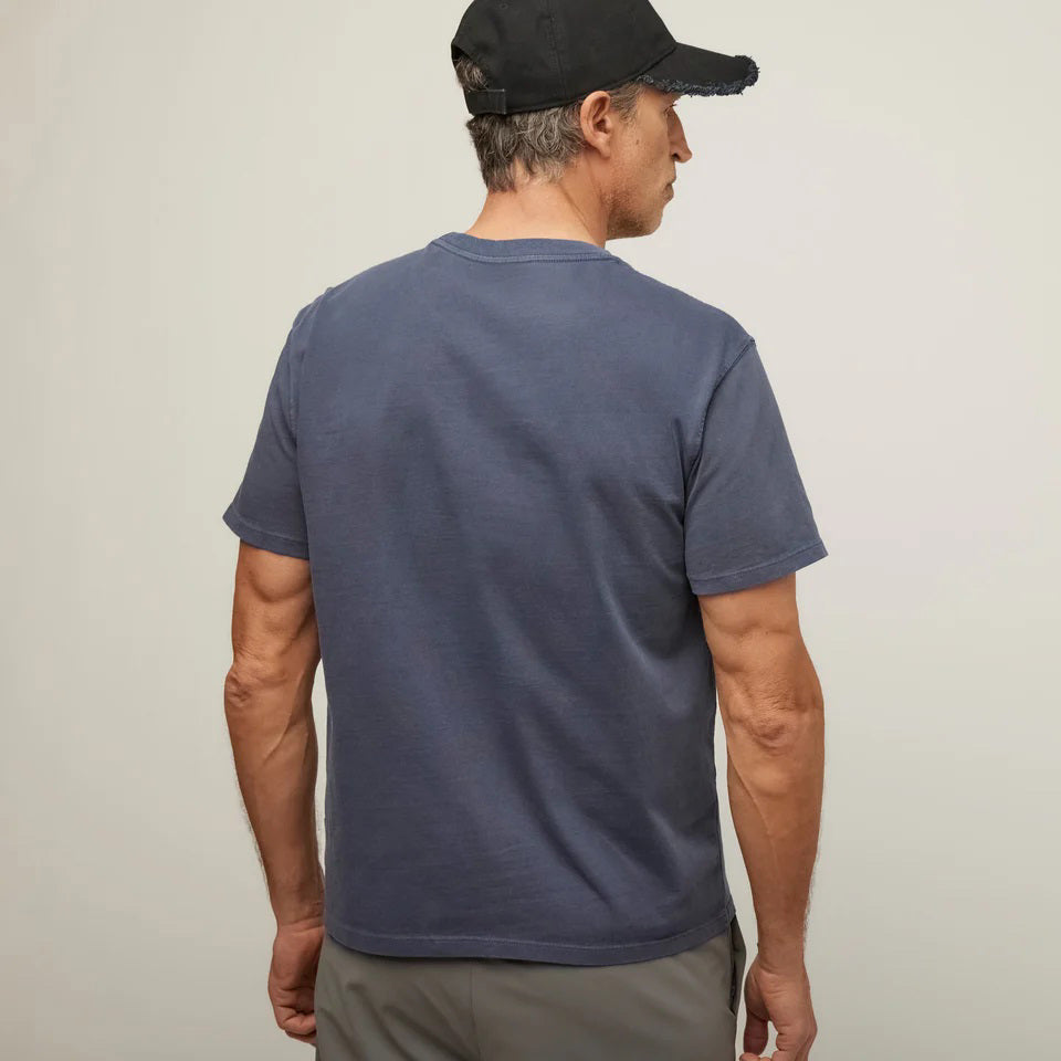 A man in a TILLEY MID WEIGHT SUNWASHED T-SHIRT IN MARINE NAVY, gray pants, and a black cap stands against a plain light background, gazing slightly to the side with a neutral expression.