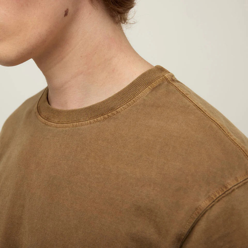 A young man in a beige bucket hat and a TILLEY Mid Weight Sunwashed T-Shirt in Peat stands against a light background, looking at the camera with a neutral expression.