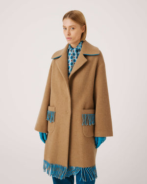 A person is wearing the SFIZIO - TWO TONED COAT WITH FRINGE by SFIZIO, featuring blue and brown fringe patch pockets and hem. They are also donning blue gloves and blue pants. The image is cropped to show from the shoulders down against a plain, off-white background.