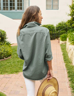 The FRANK & EILEEN - EILEEN RELAXED BUTTON UP SHIRT IN FAMOUS DENIM THYME is a light green, long-sleeved button-up shirt with a single chest pocket. The sleeves are rolled up to the elbows, and the fabric has a slightly crinkled texture. Featuring Eileen's relaxed fit, this shirt includes bust-flattering button placement, a high-low hem, and a collar without a visible tag.