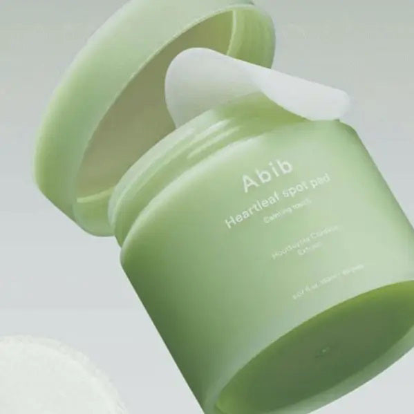 A light green jar labeled &quot;ABIB - HEARTLEAF SPOT PAD CALMING TOUCH 80 PADS&quot; from ASIAN BEAUTY WHOLESALE contains 80 pads with heartleaf extract and is shown against a plain white background.