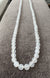 A coiled ANN LIGHTFOOT - LONG STRAND FACETED MILKY AGATE NECKLACE of translucent, round beads is displayed on a dark wooden surface. The faceted 8mm beads have a smooth and glossy finish, appearing luminous against the dark background. The necklace is attached with a sterling silver clasp.