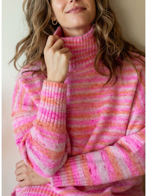 A person wearing the MERSEA - GOOD VIBRATIONS STRIPE TURTLENECK from MER SEA, a cozy sweater made of nylon, polyester, and acrylic in shades of white, gray, and beige, leans against a countertop. They have curly hair and a relaxed posture. The background includes a white vase and several stacked items.