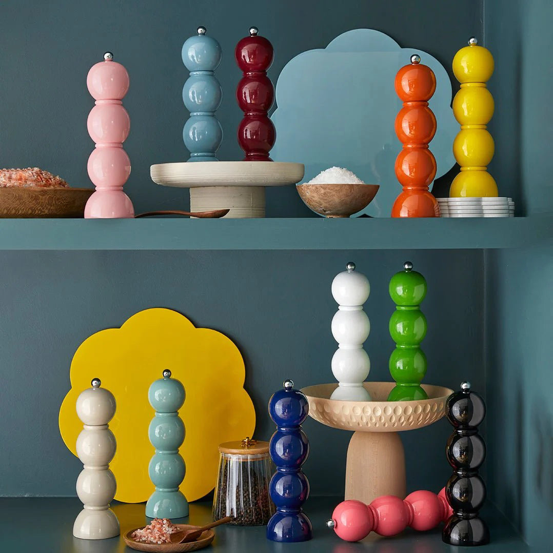 A vibrant display of ADDISON ROSS LONDON - BOBBIN SALT OR PEPPER MILL 24CM grinders in various shades such as pink, red, blue, orange, yellow, green, white, and a striking black lacquered finish. They are arranged on two shelves against a muted green background alongside various serving bowls.