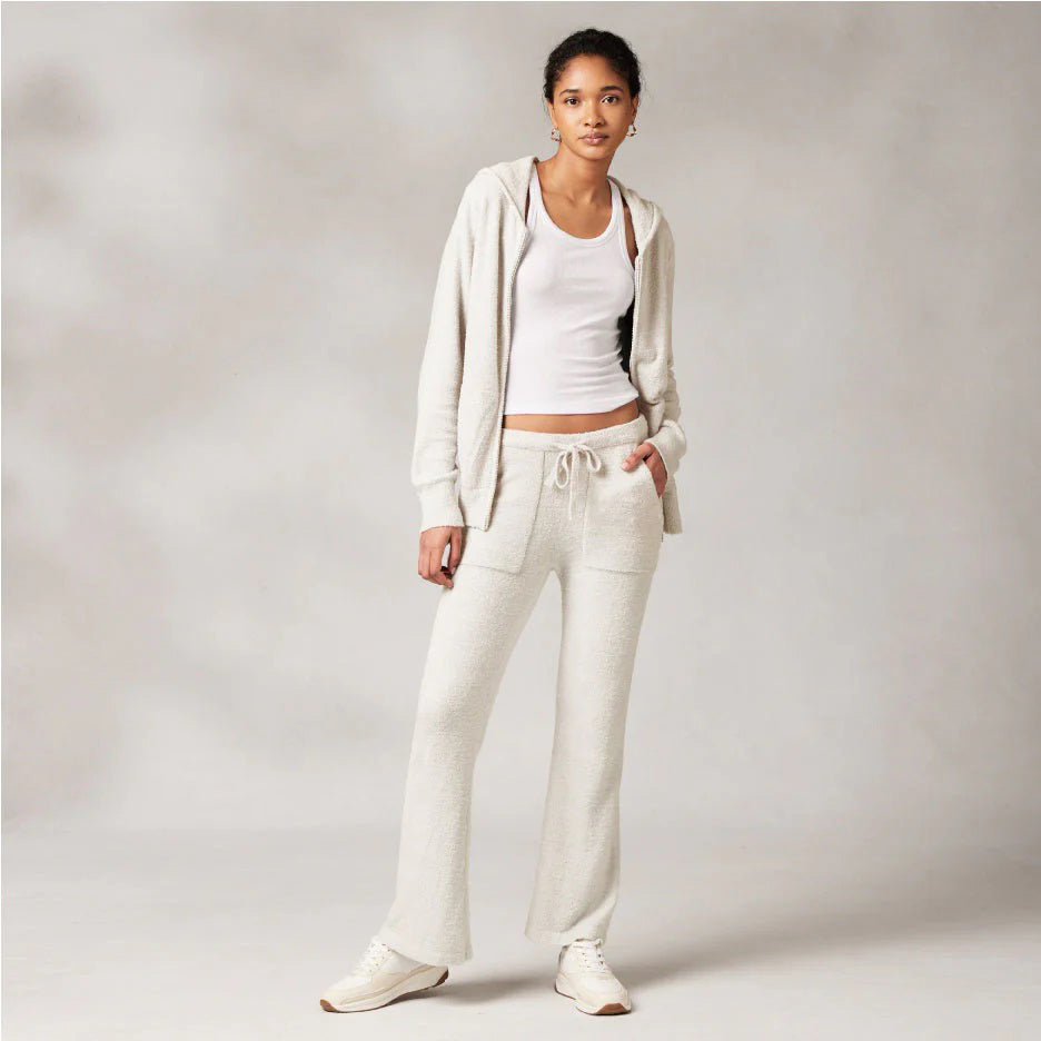 A person stands confidently against a plain background, wearing a light-colored casual outfit consisting of a zip-up hoodie, tank top, and the KASHWERE - WIDE LEG LOUNGE PANT with a drawstring waistband. They pair the outfit with sneakers and have one hand in their pocket, exuding a relaxed, stylish vibe.