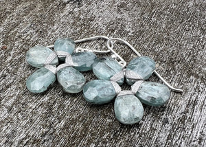 The ANN LIGHTFOOT - BLUE GREEN KYANITE FLOWER EARRINGS by ANN LIGHTFOOT, featuring faceted blue green kyanite clusters, are displayed on a weathered gray wooden surface.