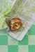 A CODY FOSTER BAGEL WITH LOX ORNAMENT, crafted from glass and detailed with sesame seeds and a green string, rests on green and white checkered paper resembling a sandwich with clear fillings. The paper showcases text and patterns in green, subtly accentuating the charm of this distinctive ornament.