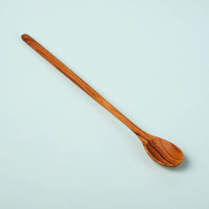 The TEAK LONG SPOON by BE HOME, INC is a handcrafted wooden utensil made in Thailand, distinguished by its round, smooth handle and shallow, oval-shaped bowl that showcase a natural wood grain pattern. Presented against a light blue background, this elegant piece is best maintained by hand washing with mild soap.