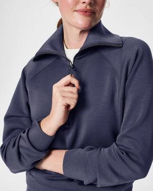 A woman with long brown hair is wearing the SPANX AIRESSENTIALS HALF ZIP sweatshirt and matching pants, both crafted from lightweight fabric for luxurious comfort. She holds her braided ponytail with one hand while looking to her right against a plain white background.