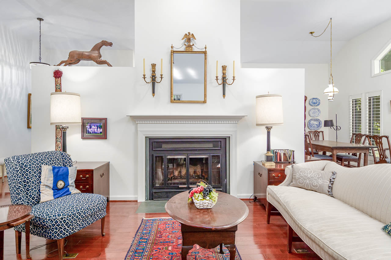 A bright, elegant living room with a fireplace, two lamps, a sofa, an armchair, a coffee table with a flower arrangement, and a dining area in the background. A large horse statue decorates the mantle.