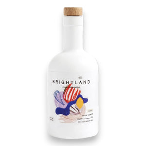 A white bottle with a wooden cap labeled "BRIGHTLAND - 'LUCID' LEMON INFUSED OLIVE OIL" features a vibrant, abstract design with shapes in red, blue, yellow, and purple. The label indicates it is made from 100% lemon-infused olive oil using early-harvest Frantoio olives sourced from small family farms. The bottle holds 375 ml or 12.7 fl oz of oil.