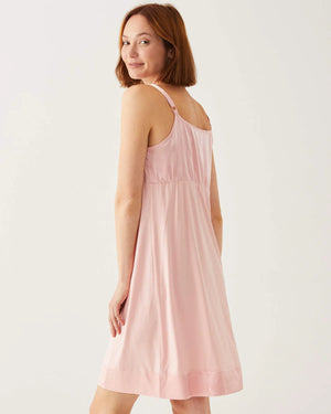 A woman with shoulder-length hair wears a light pink slip dress, like the elegant MOON DANCER PAJAMA DRESS by MER SEA, and fluffy slippers. She stands against a plain white background, looking directly at the camera with a neutral expression. Her arms are relaxed at her sides.