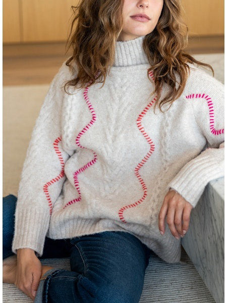 A person with long hair is wearing the MERSEA - ALPINE SWEATER by MER SEA, a loose-fitting, light grey acrylic sweater adorned with subtle purple zigzag patterns. They are also dressed in light blue jeans. The background is plain white.