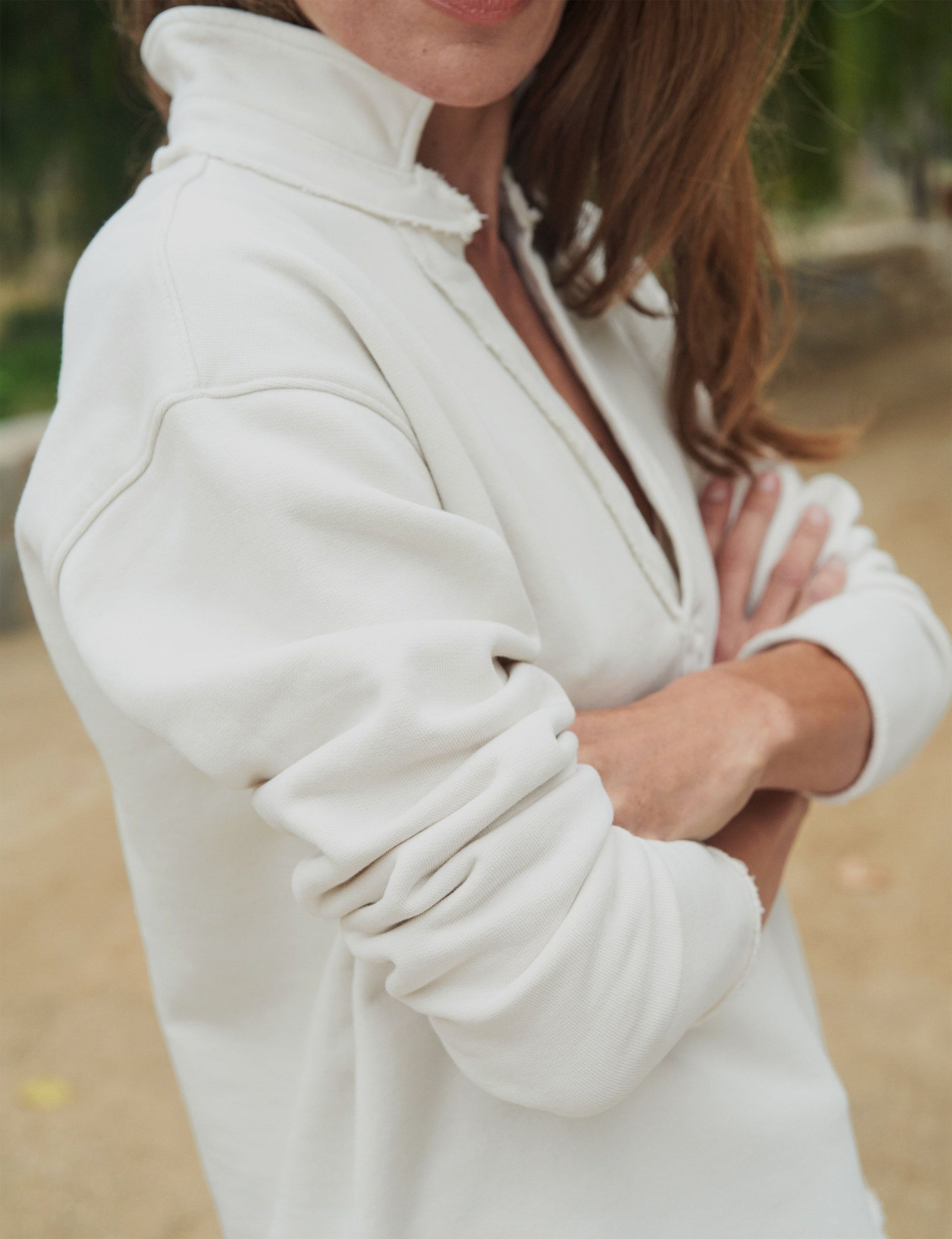 A person with long hair stands outdoors, wearing a vintage white Patrick Popover Henley in triple fleece from Frank & Eileen. Their sleeves are casually rolled up and their arms crossed, while a path and greenery can be seen in the blurred background.