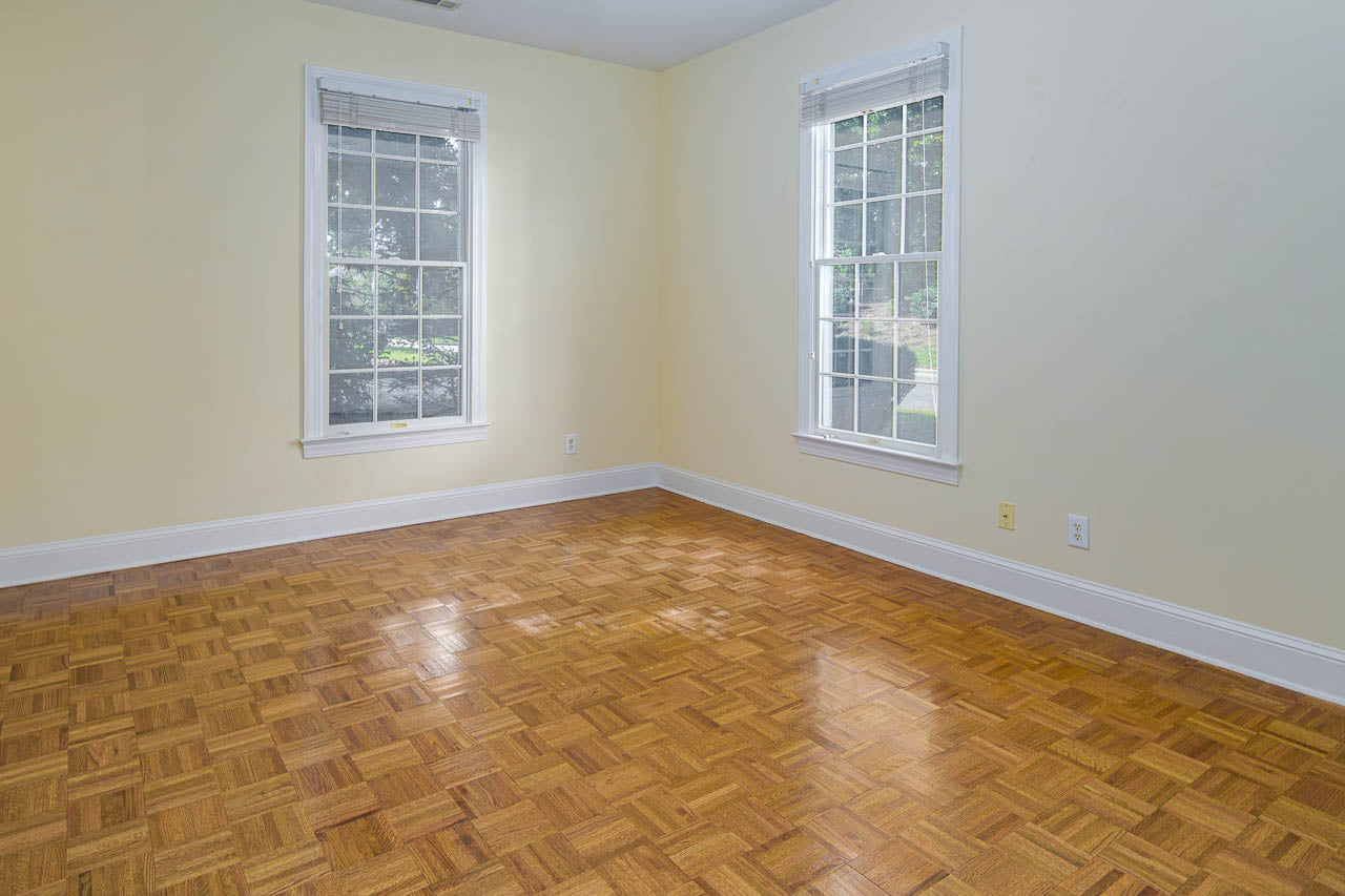 Empty room with light yellow walls, two large windows with white trim, and a polished parquet wood floor. Sunlight comes through the windows, casting reflections on the floor.