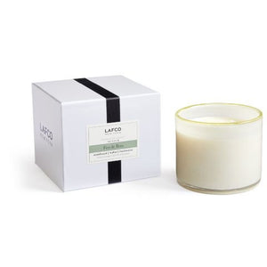 A white glass candle is next to a white and black box labeled "LAFCO New York," with a refined finish. The package hints at the earthy scent of sandalwood and features LAFCO's name prominently. .