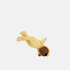 An adorable DOZY DINKUM DOLL by OLLI ELLA USA, featuring soft brown fabric skin, closed embroidered eyes, and short black hair, is dressed in a charming yellow onesie adorned with a rainbow design on the chest. Perfect as a first toy, the doll is lying on a white, quilted surface.