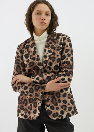 A person with wavy brown hair is wearing the ALYKI LABORATORIO LEOPARD PRINT JACKET over a white turtleneck sweater. They have their arms crossed and are standing against a plain white background.