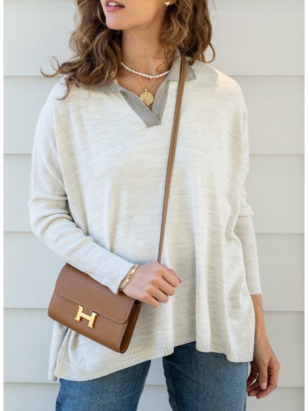 A person with wavy brown hair stands sideways against a plain white background, wearing a light-colored, loose-fitting women's sweater and blue jeans. They glance towards the camera with a neutral expression, showcasing the sporty and stylish appeal of the MERSEA - CATALINA POLO SWEATER by MER SEA.