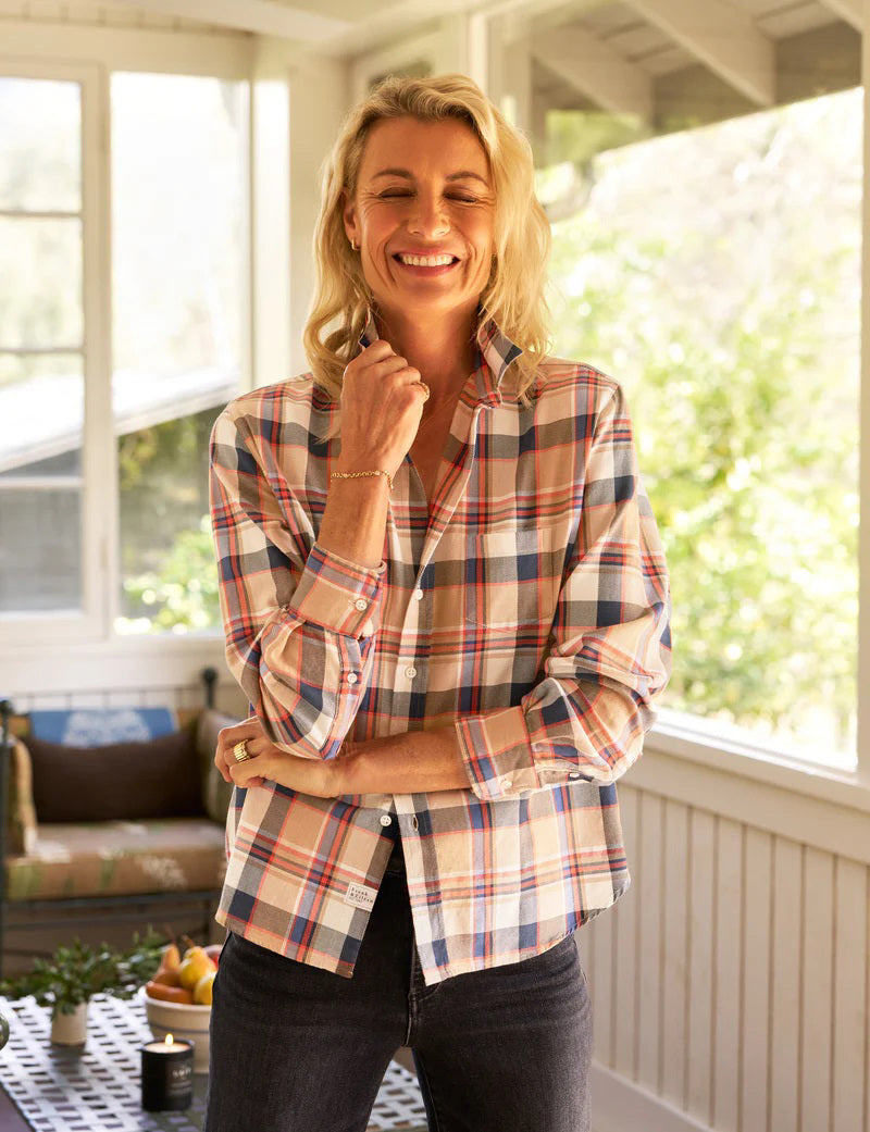 A person with long blonde hair stands in a sunlit room, facing large windows. They wear a FRANK & EILEEN Eileen Relaxed Button Up Shirt in Italian Flannel Orange Plaid, paired with dark pants. A small table with fruit is nearby, adding to the room's cozy ambiance against its white walls.