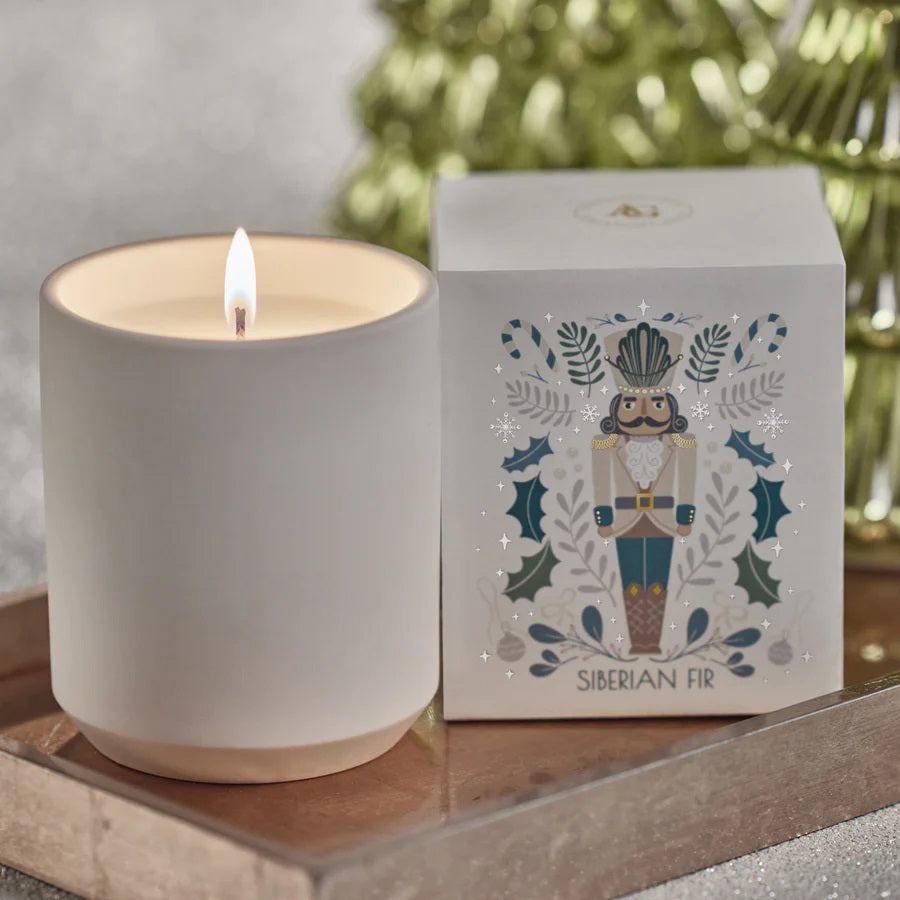 A NUTCRACKER SIBERIAN FIR CANDLE by ZODAX, housed in a minimalist white holder, is placed next to a decorative box featuring nutcracker art. Both items are elegantly arranged on a glittery tray, creating an enchanting atmosphere with fragrance notes that evoke cozy memories.