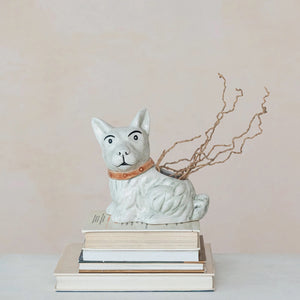 A VINTAGE REPRODUCTION DOG PLANTER by CREATIVE COOP, featuring a brown collar, is positioned atop a stack of books. Twisted branches extend from the top opening of the stoneware planter, giving it a whimsical appearance. The backdrop is a neutral beige.