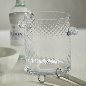 The BAGATELLE SWIRL ICE BUCKET by ZODAX is a clear glass, cylindrical ice bucket measuring 15.24 cm x 17.78 cm, adorned with subtle diagonal swirl patterns. It features two ornate looped handles on opposite sides and is showcased against a plain white background.