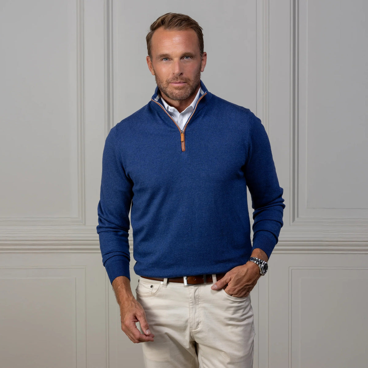 A man with light brown hair and a trimmed beard wears the COLLARS AND CO Indigo with Brown Suede Trim Cashmere Touch Australian Merino Quarter Zip Sweater over a white shirt and cream pants, standing before a light gray paneled wall.