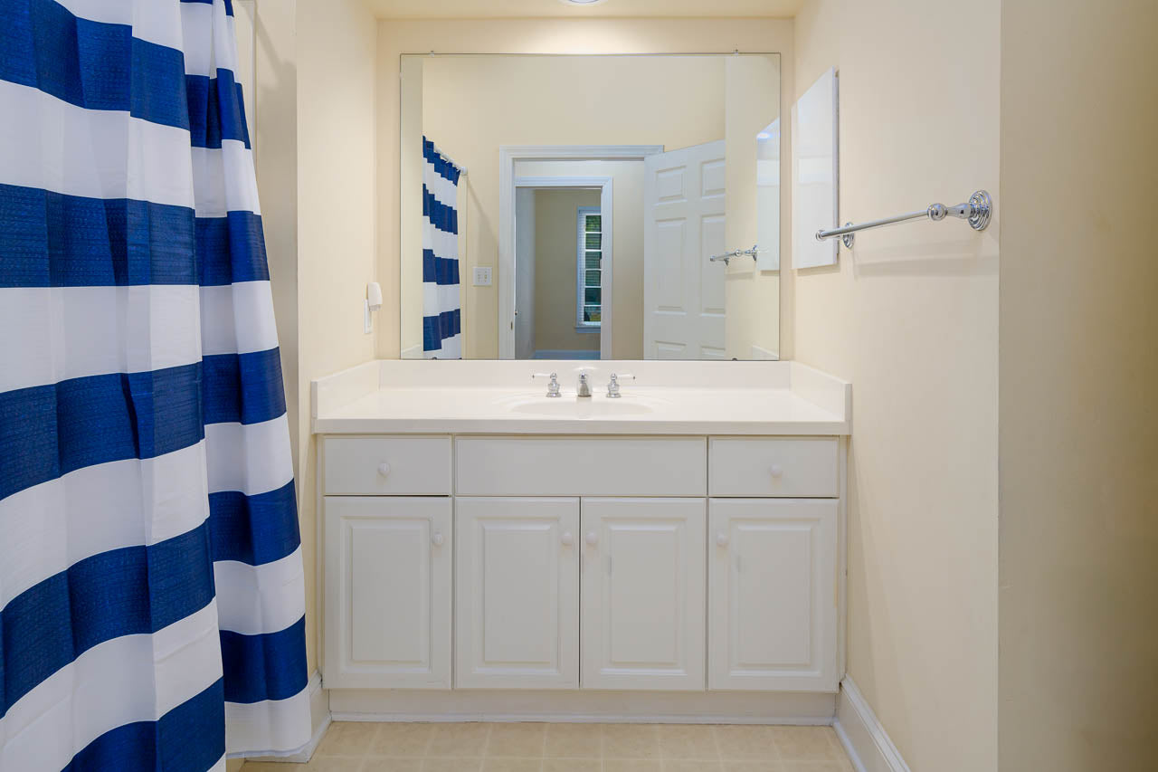 A bathroom with a white vanity, double faucets, a large mirror, and cream walls. A shower with a blue and white striped curtain is on the left, and a towel rack is on the right wall. The floor is light-colored tile.