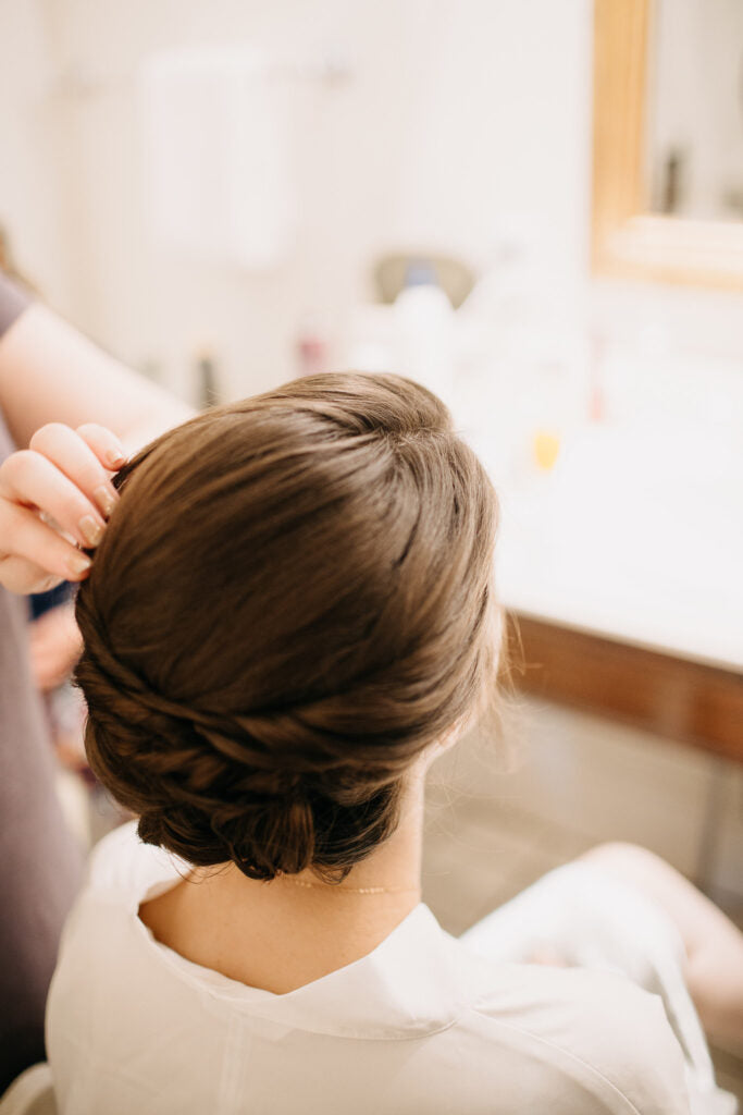 A person with brown hair styled in an elegant low updo is having their hair adjusted by another person in a softly lit room, likely a bathroom, with a blurred mirror and counter in the background.