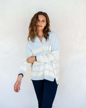 A person with shoulder-length wavy hair is standing against a light-colored wall, wearing the MERSEA - MARINA POLO SWEATER that exudes maritime spirit and dark pants. They are facing the camera with a relaxed expression as shadow patterns fall across the wall behind them.