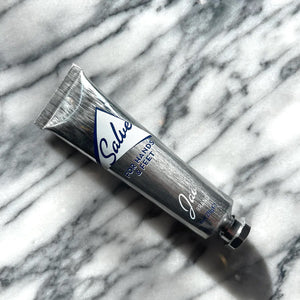 A silver tube labeled "JAO BRAND - SALVE FOR HANDS AND FEET" by JAO LIMITED, featuring essential oils, is placed inside a small white bowl. Beside the tube is a metallic golden key-like tool. The bowl sits on a marble surface.