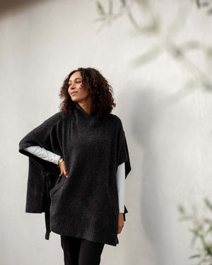 A woman with curly hair stands against a light-colored wall, wearing the MERSEA Cape Poncho Sweater from MER SEA in an oversized fit over a white long-sleeve shirt. She looks off to the side with a relaxed expression, while some blurred greenery graces the foreground.