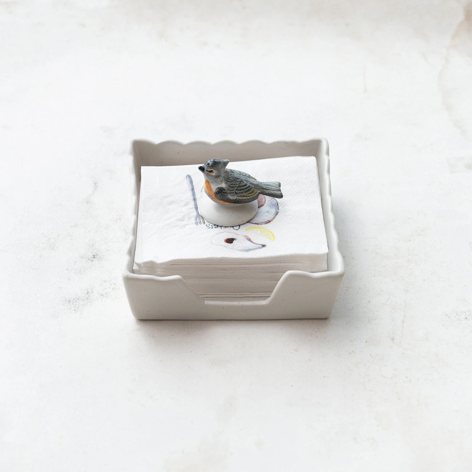 A small ceramic dish, the Creative Coop NAPKIN HOLDER WITH STONEWARE BIRD WEIGHT, contains a stack of white napkins. The top napkin features a sketch of a bird, complemented by a durable stoneware bird-shaped weight placed on the napkins. The background is a light, neutral, textured surface.