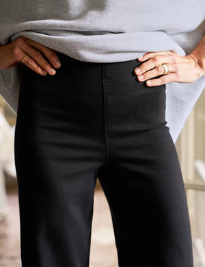 A person wearing FRANK & EILEEN Westport Illusion wide-leg pull-on jeans and black ankle boots stands in a well-lit room, with a plant on a small white table, a stack of books, and a glass door revealing a cozy living space in the background.