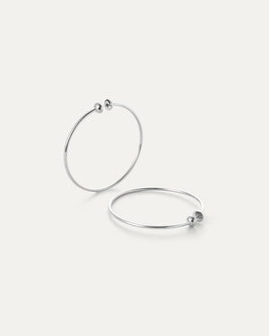 The JENNY BIRD - MEDIUM ICON HOOP EARRINGS, made from high polish stainless steel, feature two thin silver hoops with small beads at the clasp. Set against an off-white background, one earring is upright while the other lies flat.