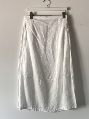 The CUT LOOSE - SIDE PLEAT SKIRT by CUT LOOSE hangs on a hanger against a plain grey background. The long white maxi skirt appears to be made of lightweight, slightly wrinkled linen fabric, with a simple design free of patterns or embellishments.