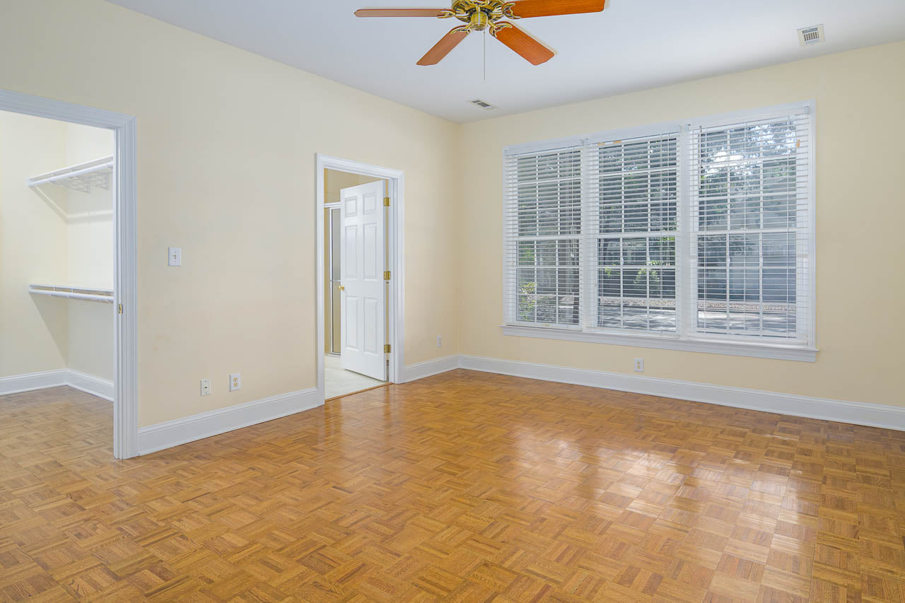 Empty room with light yellow walls, parquet wood flooring, a ceiling fan, large window with white blinds, a door leading to a walk-in closet, and another door to a bathroom. Natural light fills the space.