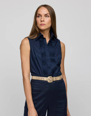 A woman with long hair wears the HINSON WU Lilith Sleeveless Shirt, made from stretch polished cotton with pleats and a beige belt, against a gray background. She exudes confidence as she gazes at the camera, highlighting the shirt's versatility.