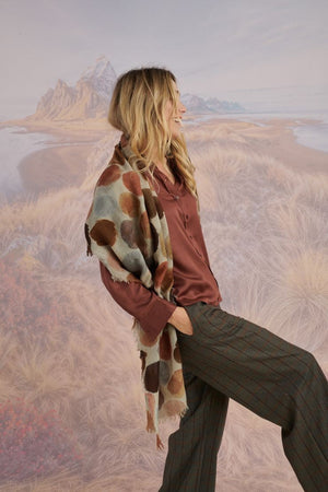 A woman with blonde hair displays the STORIATIPIC - BETTY WOOL SCARF, a large rectangular wool accessory with earth-toned polka dots, set against a scenic mountain landscape.