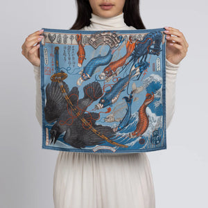 A person is wearing a white turtleneck and holding the SABINA SAVAGE - KOINOBORI KARAUS SILK TWILL SCARF 42CM by SABINA SAVAGE ENGLAND, a vibrant piece featuring dynamic koi fish and Hokusai-inspired waves on a blue background, complete with intricate Edo-period Japanese patterns.