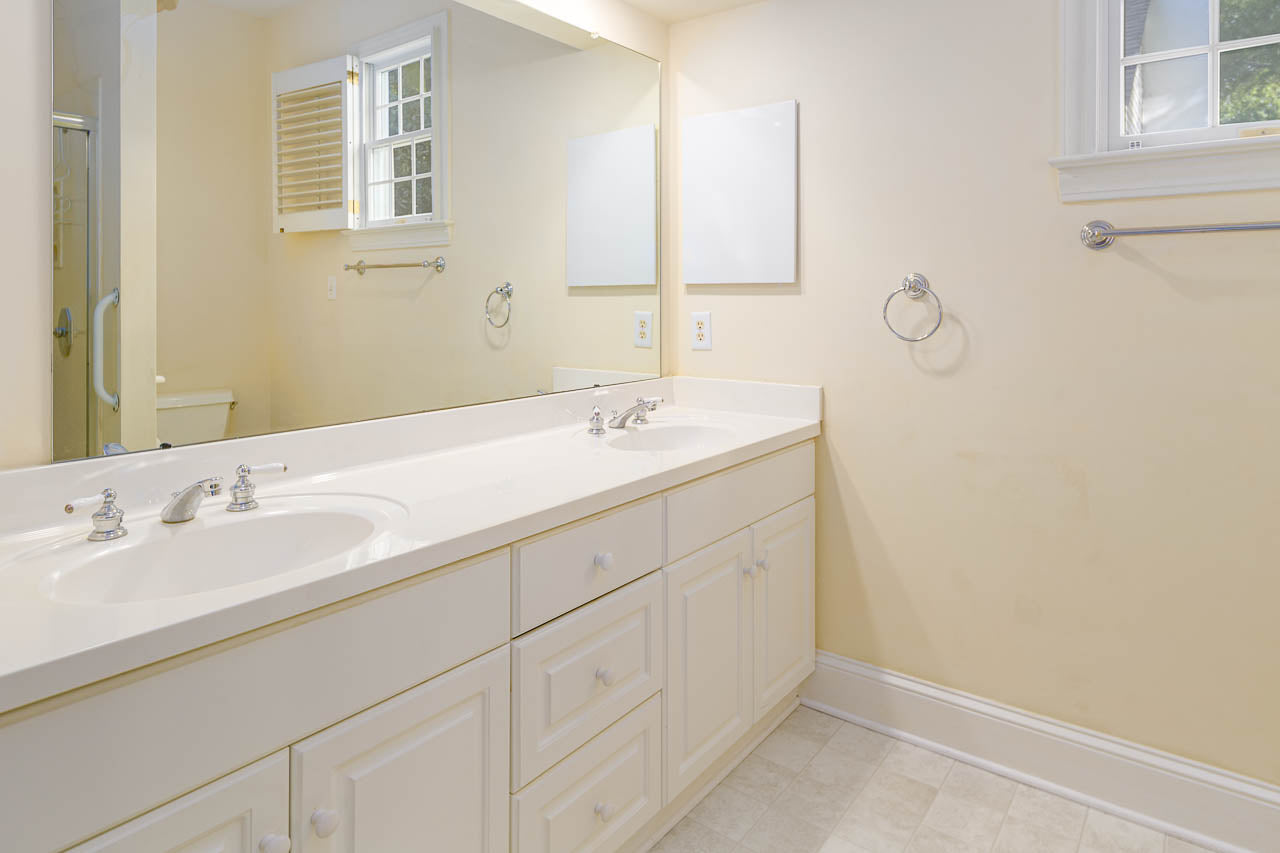 A clean, bright bathroom with a double-sink white vanity, white cabinets, large mirror, towel rings, and windows letting in natural light. The walls and floor are light-colored.
