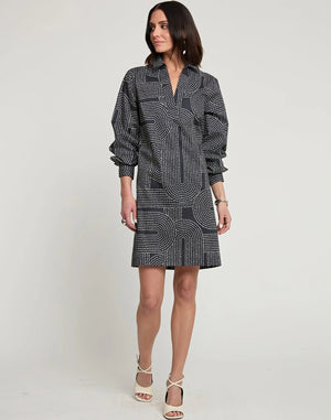 A woman with shoulder-length dark hair is wearing the new HINSON WU Vicky Long Sleeve Dress with Saddle Stitch Print, a knee-length outfit featuring a geometric dot pattern in shades of grey and white. Paired with white high-heeled sandals, she stands against a plain, off-white background.