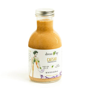 A 10 fl oz (295 ml) bottle of Dress It Up Dressing - Caesar dressing. The label showcases an illustration of a person in a green dress and emphasizes the product's low-calorie formulation made with olive oil and chia seeds, highlighting its simple, wholesome ingredients. The bottle is topped with a metal screw cap.