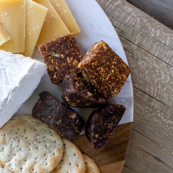 A close-up of a charcuterie board featuring a variety of foods. On the left, there are slices of yellow cheese and creamy white cheese. In the middle are several pieces of FICHI - FIG AND WALNUT BITES from FICHI. At the bottom, there are round crackers with poppy seeds—a nutritious snack for any occasion.