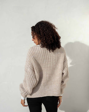 A woman with curly hair stands against a light-colored wall, wearing the MERSEA - BEACHSIDE TURTLENECK from MER SEA over a white shirt, black pants, and black flat shoes. Beside her is a woven cylindrical ottoman. She gazes thoughtfully to the side with a relaxed posture.