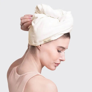 The QUICK DRY HAIR TOWEL by KITSCH is packaged in a cream-colored box, detailed with a black droplet shape and an inset image of someone wearing the highly absorbent towel. Designed for fine, curly, or straight hair, it's crafted from organic cotton and bamboo. The text and icons on the packaging are clearly visible.