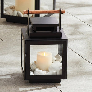 The ADWIN OUTDOOR LANTERN by NAPA HOME AND GARDEN is a black metal lantern with glass panels and a teak handle on top. Inside, there's a white pillar candle. The design is modern and exclusive, featuring clean lines and a sturdy structure, making it an ideal choice for both indoor and outdoor use.