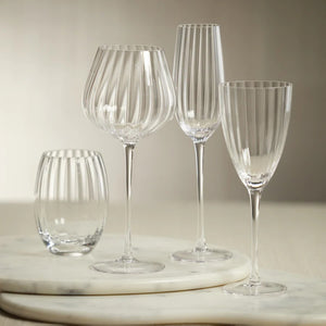 The MADELEINE OPTIC CHAMPAGNE FLUTE by ZODAX is a tall and slender clear crystal glass adorned with vertical grooves extending from the rim to the stem, which rests on a flat, round base. The empty flute showcases a polished, reflective finish that elevates its elegance, making it perfect for special occasions and toasting celebrations.